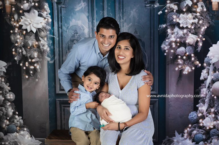 A family laughs and hugs in front of a blue door, decorated with a wreath and lights. Avnida Photography captures the joy of the season in this holiday photo.