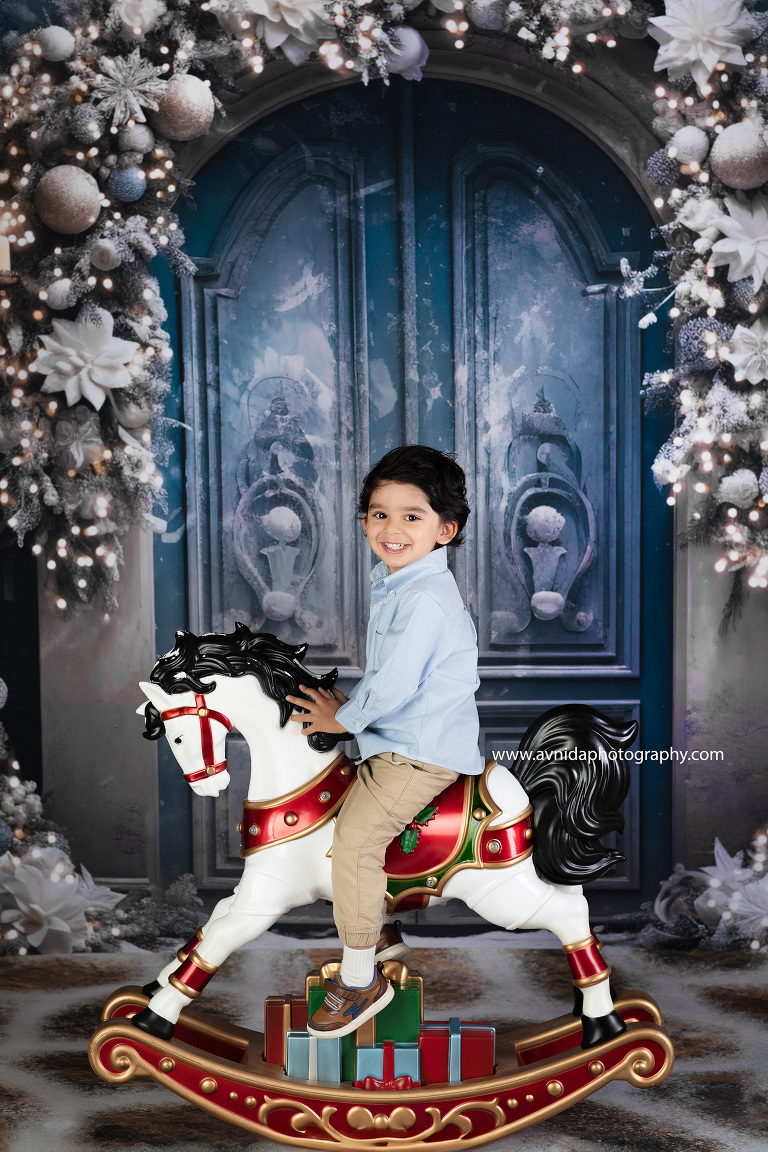 A child's laughter fills the air as they gleefully ride a horse, framed against a charming blue door adorned with festive decorations. Avnida Photography captures the magic of the holidays with this heartwarming and joyful portrait.